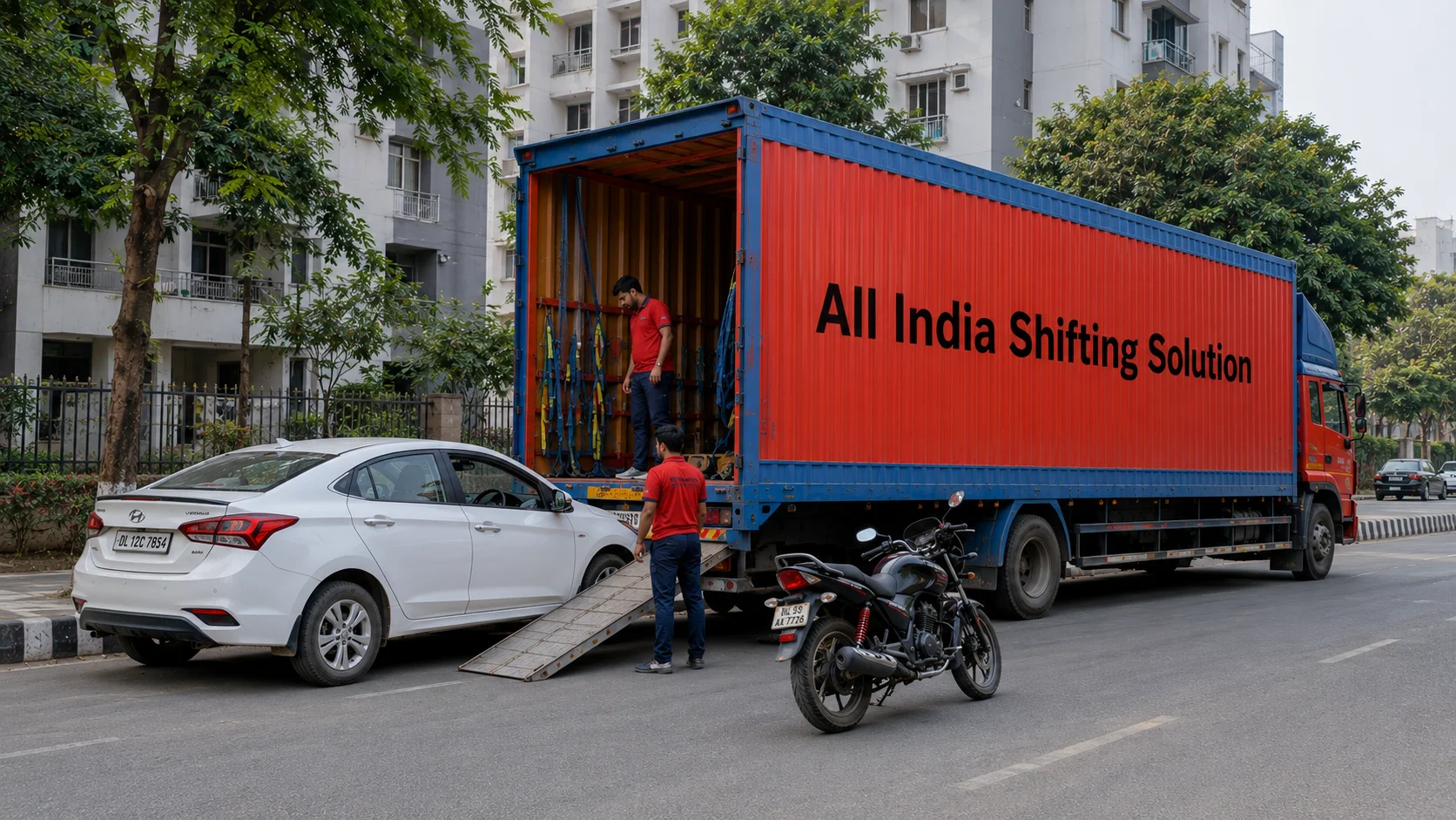 A wide shot of a massive enclosed a plain red and blue Indian container truck with "All India Shifting Solution" in large black text parked on a wide Indian urban road, with both a car and a bike staged safely for loading.